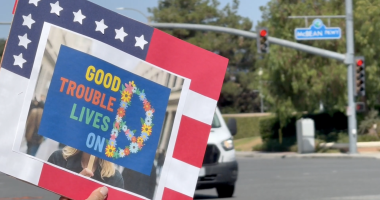 A person holds a sign at a street intersection with the phrase “GOOD TROUBLE LIVES ON” written in colorful letters next to a peace sign made of flowers. The sign is bordered in red, white, and blue with stars, resembling the American flag. In the background, a street sign reads “McBean Pkwy” and a white van waits at a red light.